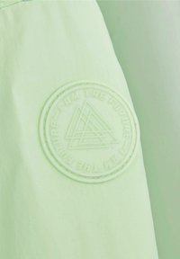 Hellgrüner Stoff mit glatter Textur zeigt ein kreisförmiges geprägtes Patch mit der Aufschrift "ICH BIN DIE ZUKUNFT" und einem geometrischen Design.