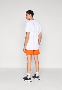 Wit T-shirt met korte mouwen en een geribbelde textuur, gecombineerd met feloranje korte broek, gedragen door een persoon met de rug naar de camera, die zwarte sneakers draagt.