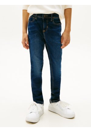 Kind trägt dunkelblaue, eng anliegende Jeans und weiße Turnschuhe, steht vor einem schlichten hellen Hintergrund.