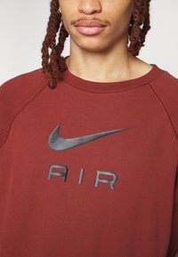 Djupröd sweatshirt i mjukt tyg, med en texturerad grå Nike-logga och ordet "AIR" i fet stil över bröstet.