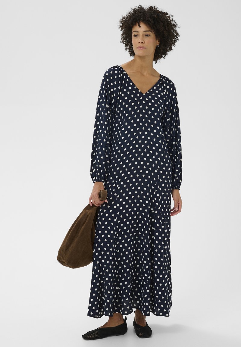Robe maxi bleu marine à pois avec manches longues et col en V. Dotée d'une jupe fluide, accompagnée d'un sac à main marron et de ballerines noires.