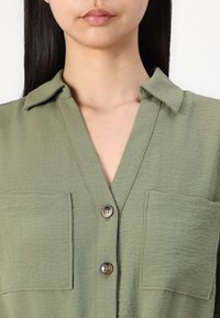 Chemise vert à manches longues avec col, deux poches poitrine, tissu texturé et décolleté en V. Présente une fermeture à boutons sur le devant.