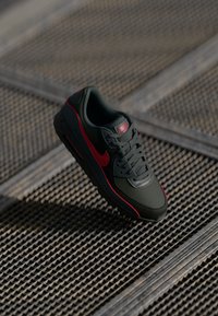 Crne i crvene Nike Air Max tenisice vise iznad pozadine od teksturirane metalne rešetke.