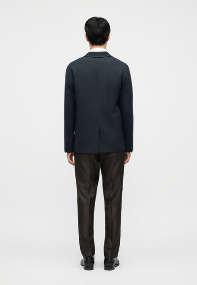 Μπλε σκούρο σακάκι με κομμένη φόρμα και δομημένη πλάτη, σε συνδυασμό με καφέ καρό παντελόνια και απλά μαύρα παπούτσια. Καθαρές γραμμές και κλασικός σχεδιασμός.