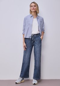 Kobieta stojąca na jednolitym tle, ubrana w pasiastą koszulę zapinaną na guziki, białą koszulkę, szerokie dżinsy z paskiem do wiązania oraz białe trampki.