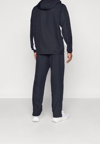 Mann von hinten gesehen, der eine schwarze Kapuzenjacke mit weißen Streifen an den Ärmeln, schwarze Hosen und weiße Sneakers trägt, vor einem einfarbigen Hintergrund steht.