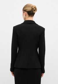 Blazer nero su misura con una vestibilità strutturata, revers a punta e bottoni decorativi sulle maniche. Tessuto liscio con una silhouette elegante.