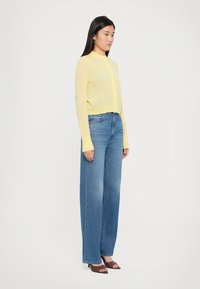 Lys gul cardigan med knapper, parret med løse blå denimjeans. Modellen har brune hæle sandaler på. Enkel design og afslappet pasform.