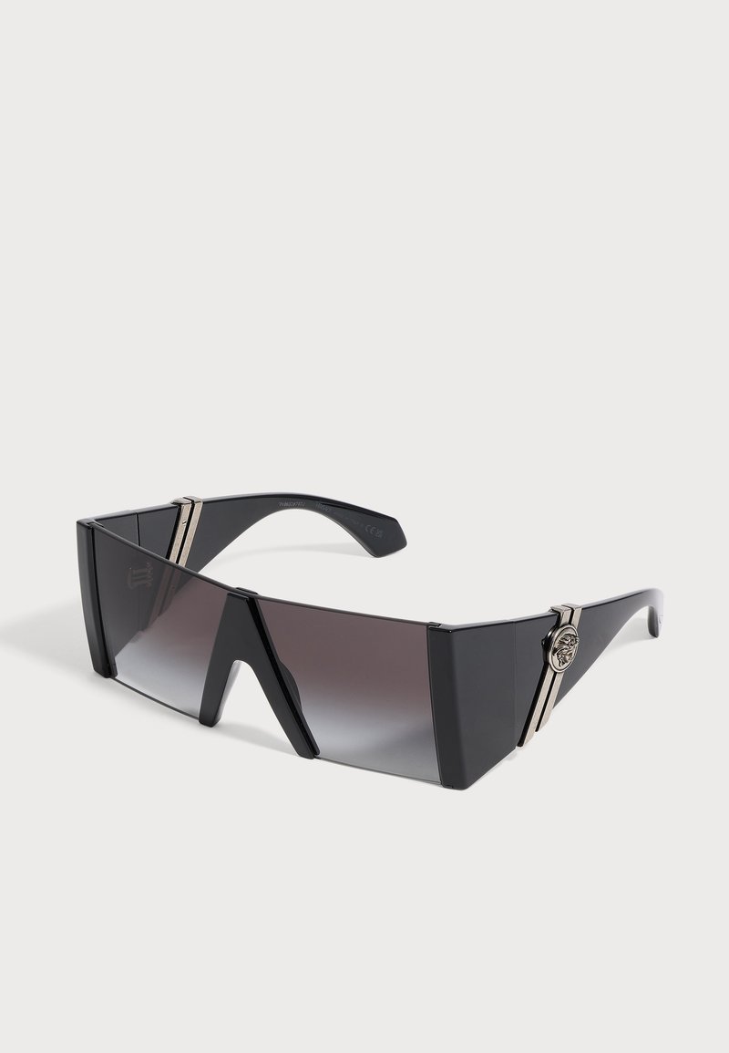 Lunettes de soleil noires surdimensionnées en forme de bouclier avec verres dégradés foncés et accents argentés sur les branches sur fond blanc.