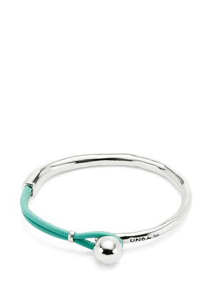 Brazalete de plata con un diseño suave y redondeado, que presenta una banda de cuero turquesa y un acento de bola de plata. Estilo minimalista.
