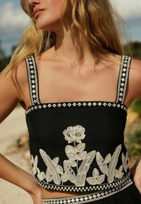 Top corto negro con escote cuadrado, que presenta bordados florales blancos en el dobladillo y tirantes con patrones intrincados. Tejido texturizado.