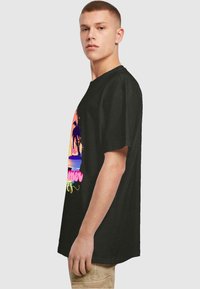 Zwart T-shirt met een relaxte fit, voorzien van een kleurrijke zonsonderganggrafiek met palmbomen en de tekst "Zomer" in roze- en geeltinten.