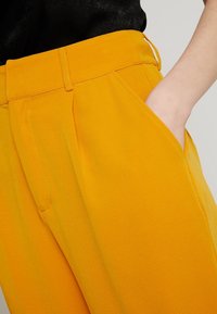 Světlé žluté oblekové kalhoty s hladkým povrchem, charakterizované rovný střih, bočními kapsami a úhlednými sklady v pase.