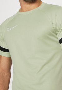 Hombre con camiseta de Nike verde claro con el logo blanco en forma de swoosh y detalles en franjas negras en las mangas, visto desde la mitad del pecho hasta el cuello.