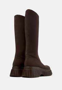 Botas de punto marrón oscuro y altas con suelas de goma gruesas y estriadas, tacones ligeramente inclinados y una textura superior suave y sin costuras.