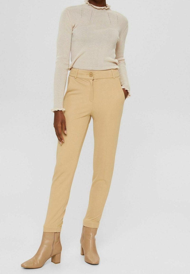 Nainen, jolla on beigehousut, luonnonvalkoinen, reunuksilla varustettu pitkähihainen ribbineuletoppi ja ruskeat nilkkurit korkokengillä, seisomassa neutraalin taustan edessä.