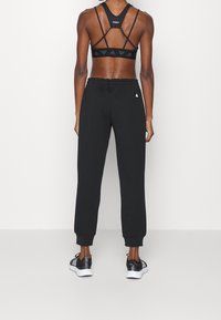 Černý sportovní outfit zahrnující sportovní podprsenku s křížovými ramínky a vysokými kalhotami s manžetami na kotnících, doplněný černými teniskami.