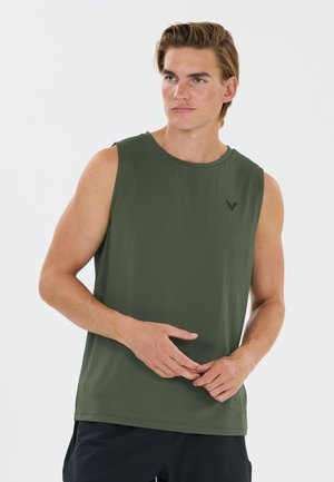 Ung mand i en ærmeløs olivengrøn sports-top og sorte shorts, der kigger til siden med hænderne løst foldet foran.