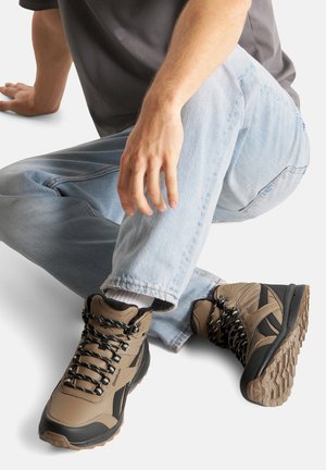 Brūni pārgājienu zābaki ar melniem akcentiem, ar teksturētu virsmu, sarežģītu šņorēšanas sistēmu un izturīgu gumijas zoli.