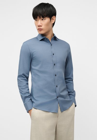 Άνδρας με μαύρα μαλλιά, ντυμένος με στενή γαλάζια πουκάμισο με κουμπιά και μπεζ παντελόνι, κοιτάζει προς τα αριστερά με τα χέρια στις τσέπες.