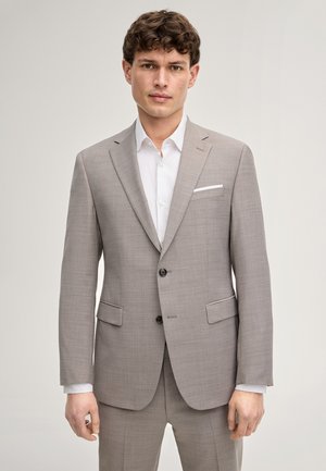 Junger Mann mit braunen lockigen Haaren trägt einen hellgrauen Anzug und ein weißes Hemd, steht vor einem schlichten hellen Hintergrund.