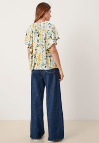 Blumige Bluse mit gelben, blauen und schwarzen Mustern, mit kurzen Rüscheärmeln und gerafftem Ausschnitt, kombiniert mit dunkelblauben weit geschnittenen Jeans.