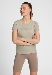 Helle beige T-Shirt mit dem "HUMMEL"-Logo in dunklerer Schrift, kombiniert mit figurbetonten braunen Radshorts, die ein vertikales Logo an der Seite haben.