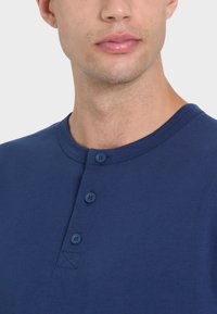 Maglietta henley in cotone blu navy con scollatura rotonda, placca con tre bottoni e un piccolo dettaglio ricamato sul petto.