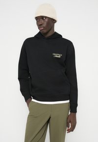 Άνδρας με μαύρο φούτερ με κουκούλα με το κείμενο «Originals Studio», μπεζ σκούφο, ελαιδόχρωμο παντελόνι, στέκεται μπροστά σε λευκό φόντο με τα χέρια στις τσέπες.