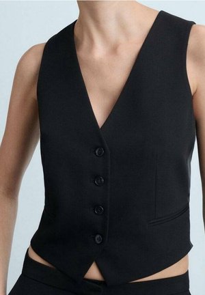 Personne portant un gilet noir cintré boutonné avec un col en V et un design sans manches, associé à un pantalon noir sur un fond clair.