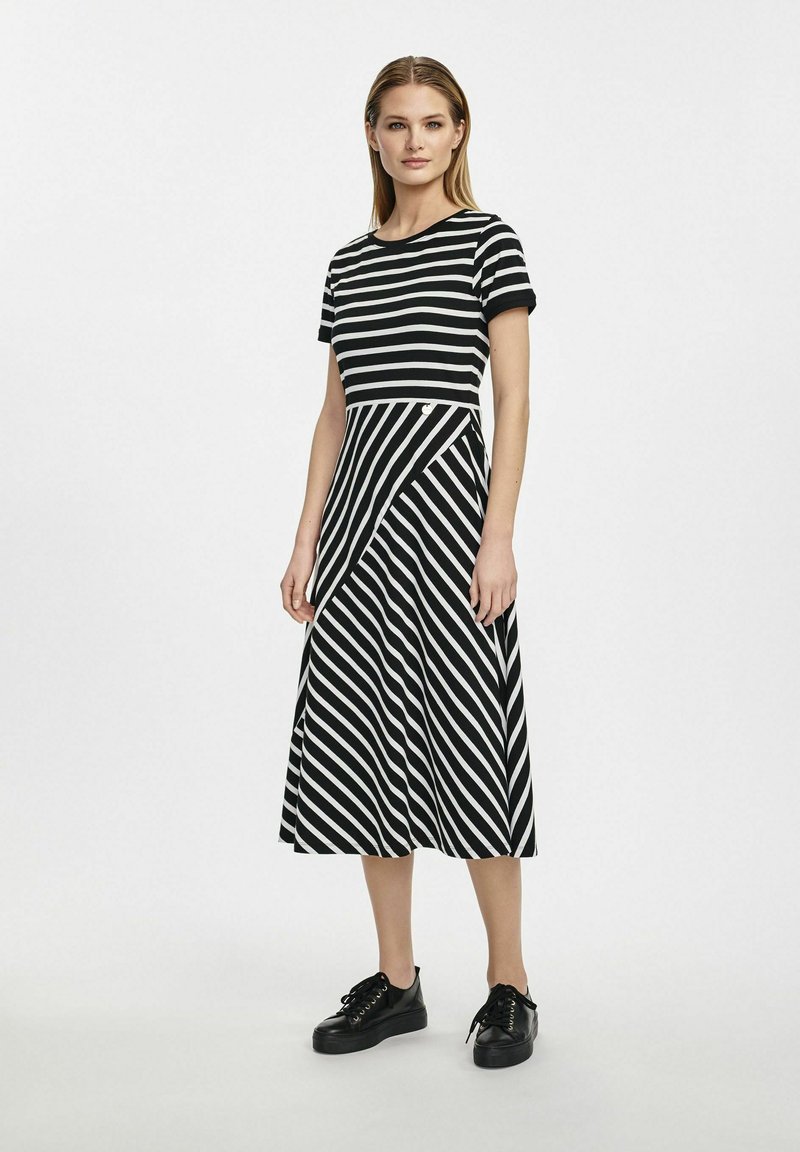 Czarna i biała sukienka w paski z okrągłym dekoltem, krótkimi rękawami i asymetrycznym dołem. Noszona z czarnymi trampkami na platformie.