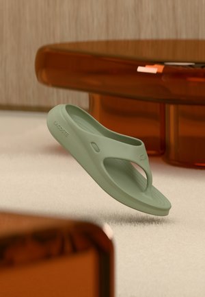 Tongs en caoutchouc vert clair avec une semelle texturée, présentant un design découpé et un logo en relief sur le côté, flottant au-dessus d'une surface douce.