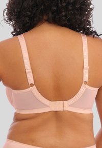 Donna che indossa un reggiseno rosa chiaro con spalline regolabili e chiusura a gancio, vista di spalle su uno sfondo neutro.