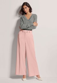 Lichtgrijze trui met V-hals, gecombineerd met wijde roze broek met knoopsluiting en persplooien. Witte hoge hakken.