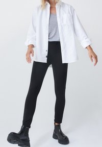 Bílá košile s knoflíky přes šedý top, doplněná černými legínami a robustními černými botami; košile má kapsu a hladkou texturu.