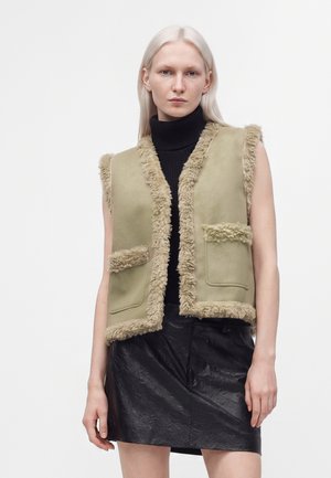 Femme aux longs cheveux clairs portant un gilet beige en shearling par-dessus un col roulé noir et une jupe noire texturée sur un fond uni.