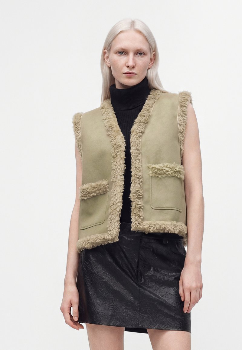 Femme aux longs cheveux clairs portant un gilet beige en shearling par-dessus un col roulé noir et une jupe noire texturée sur un fond uni.