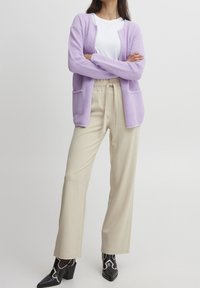 Lavendelstickad cardigan, vit T-shirt med rund hals, beige avslappnade byxor och svarta klackskor med silverdetaljer. Texturerat tyg och fickor.