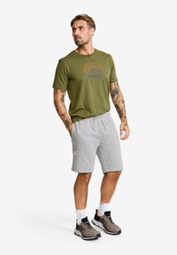 Olivgrön t-shirt med grafiskt tryck, grå shorts, grå sneakers med svarta och gula accenter, atletisk passform, avslappnad stil.