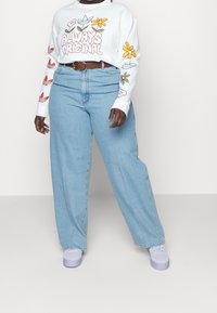 Vit långärmad skjorta med blommönster och texten "ALLTID ORIGINAL" som matchas med högmidjade ljusblå vida jeans och vita sneakers.