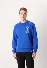 Blauwe sweatshirt met een ronde hals en ribgebreide manchetten, met een witte grafische print op de borst. Geselecteerd met zwarte broeken.