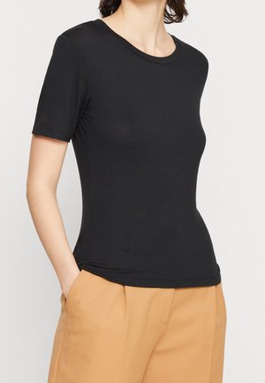 Woman wearing a fitted black short-sleeve T-shirt tucked slightly into high-waisted beige pants with hands in pockets.