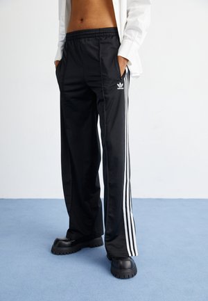 Person iført sorte Adidas træningsbukser med hvide striber og sorte chunky sko, stående på blå gulv, hænderne i lommerne.