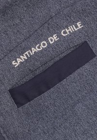 Grijze stoffen zak met marineblauwe voering, bevat geborduurde tekst "SANTIAGO DE CHILE" in het wit. Tekst gecentreerd nabij de bovenkant van de zak.