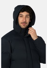 Donkerblauwe gewatteerde jas met capuchon, voorzien van een hoge kraag, rits aan de voorkant en drukknopen. Het materiaal lijkt glad en geïsoleerd.