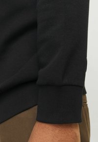 Czarny sweter z żebrowanym mankietem, ukazujący teksturowany materiał. Ręka jest ustawiona pod kątem w stosunku do khaki spodni.