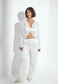Weißes Spitzencrop-Top mit Bindedetail, longärmlig und mit tiefem V-Ausschnitt, kombiniert mit weit geschnittenen weißen Satin-Hosen. Strukturierter Stoff.