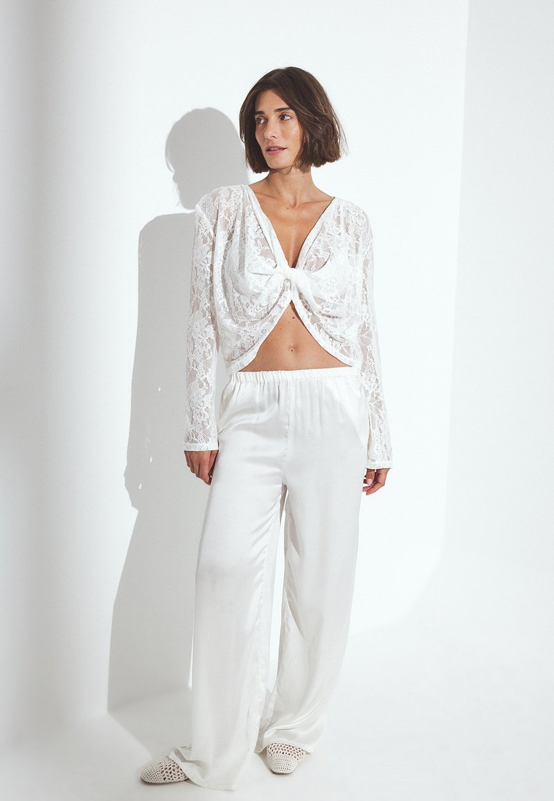 Weißes Spitzencrop-Top mit Bindedetail, longärmlig und mit tiefem V-Ausschnitt, kombiniert mit weit geschnittenen weißen Satin-Hosen. Strukturierter Stoff.