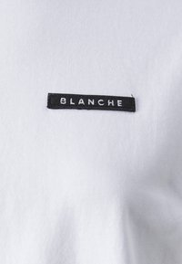 White cotton t-shirt featuring a rectangular black label with the word "BLANCHE" embroidered in white. Smooth texture, casual design.