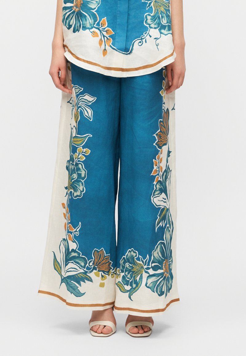 Μπλε παντελόνια φαρδιά με μεγάλα λευκά λουλουδέ νια σχέδια περιμετρικά από μπεζ ρίγες, συνδυασμένα με λευκά σανδάλια ανοιχτής μύτης.